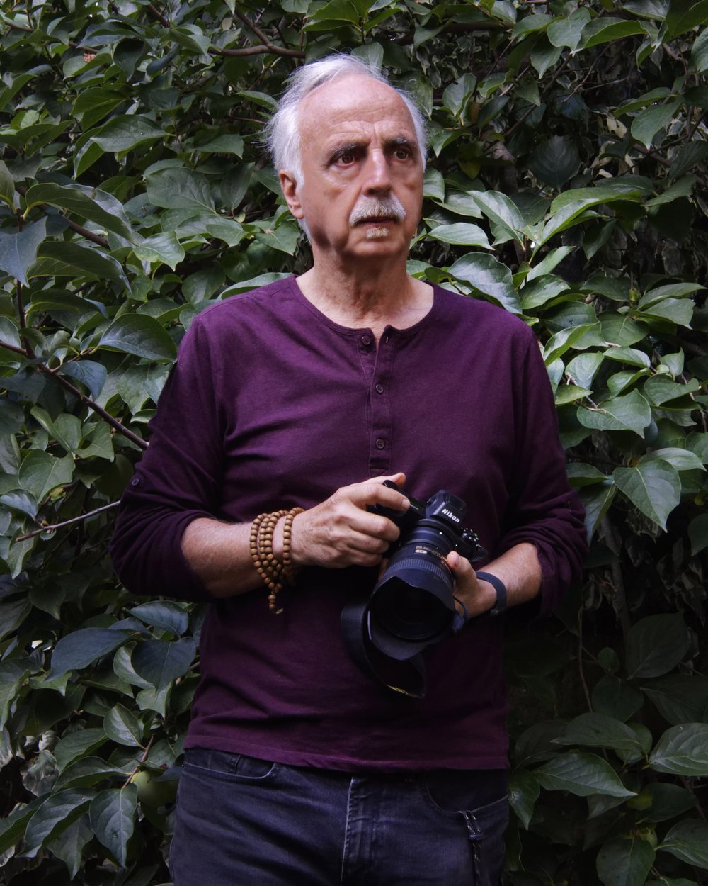 Andrea Pistolesi, holding a camera, standing against dense foliage.